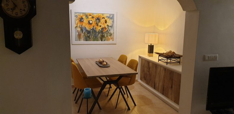Huiselijke eetruimte met houten tafel, gele stoelen en een schilderij van zonnebloemen.